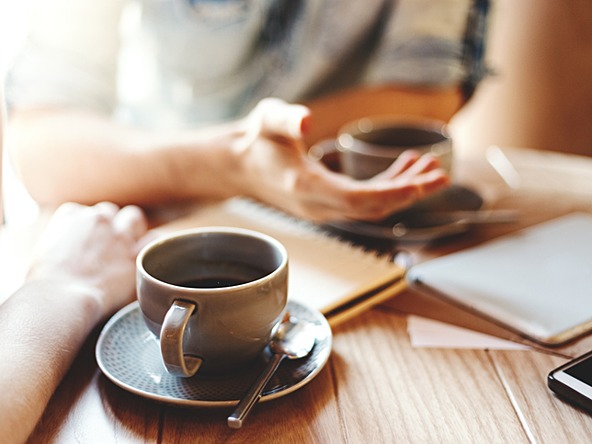 two people having cups of coffee while moving hands, with notebooks and phone